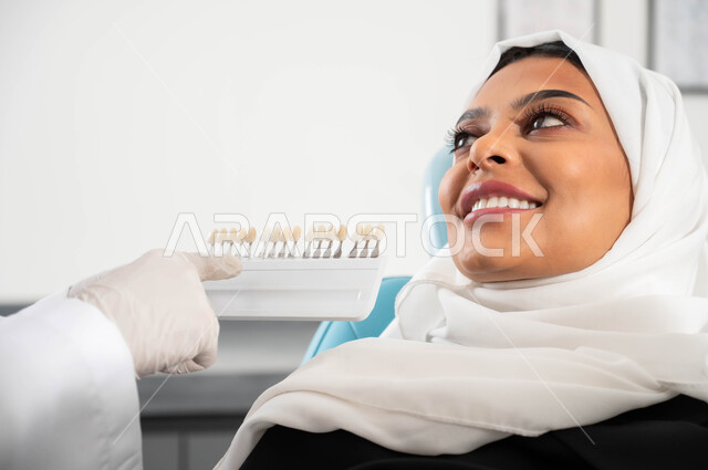 سيدة عربية خليجية سعودية محجبة مبتسمة في عيادة سنية تتلقى العلاج اللازم لصنع ابتسامة مميزة، صورة مقربة لأيدي طبيب أسنان يرتدي القفازات الطبية يمسك بيده دليل الألوان لترميم أسنان المريضة، مفهوم الرعاية والاهتمام بصحة ومظهر الأسنان
