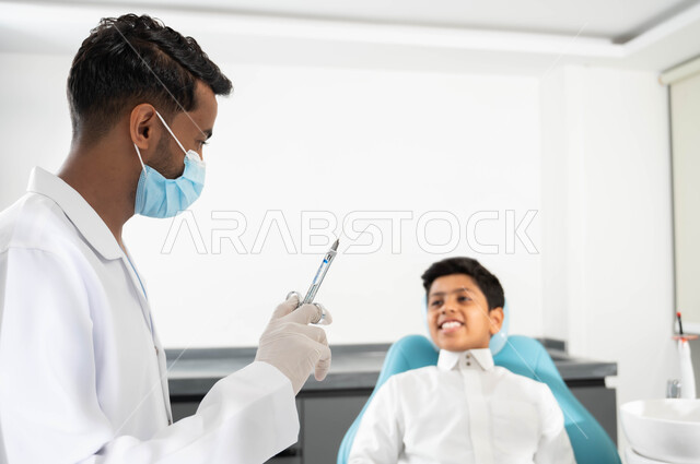 تهيأة البنج الموضعي لتخفيف الألم، مفهوم حفر السن وإزالة التسوس، ولد عربي خليجي سعودي يرتدي الثوب التقليدي يجلس في العيادة السنية يتلقى العلاج اللازم، طبيب أسنان يرتدي القفازات والكمامة الطبية يمسك الإبرة، مراجعة العيادات الصحية