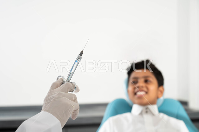 تهيأة البنج الموضعي لتخفيف الألم، مفهوم حفر السن وإزالة التسوس، ولد عربي خليجي سعودي يرتدي الثوب التقليدي يجلس في العيادة السنية يتلقى العلاج اللازم، صورة مقربة ليد طبيب أسنان يرتدي القفازات الطبية يمسك الإبرة، مراجعة العيادات الصحية