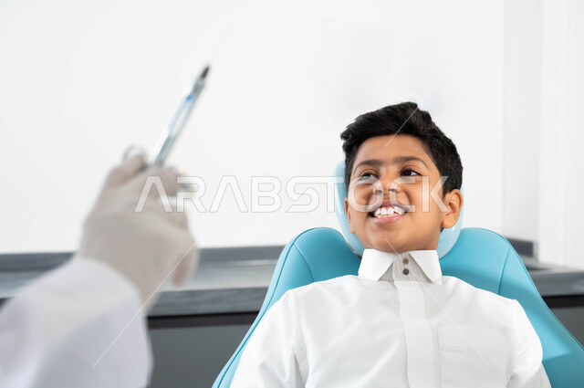 تهيأة البنج الموضعي لتخفيف الألم، مفهوم حفر السن وإزالة التسوس، ولد عربي خليجي سعودي يرتدي الثوب التقليدي يجلس في العيادة السنية يتلقى العلاج اللازم، صورة مقربة ليد طبيب أسنان يرتدي القفازات الطبية يمسك الإبرة، مراجعة العيادات الصحية