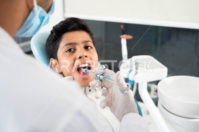 طبيب أسنان عربي خليجي سعودي يرتدي المعطف والقفازات والكمامة الطبية يقوم بحقن البنج الموضعي لتخفيف الألم، ولد يجلس في العيادة السنية يتلقى العلاج اللازم، مفهوم حفر السن وإزالة التسوس، زيارة العيادات الصحية واتخاذ الإجراءات اللازمة