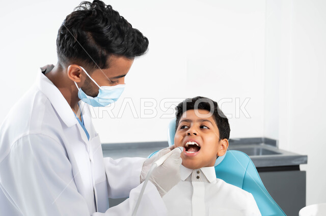 ولد يجلس في العيادة السنية يتلقى العلاج اللازم، طبيب أسنان عربي خليجي سعودي يرتدي المعطف والقفازات والكمامة الطبية يمسك بيده أداة حفر السن وإزالة التسوس، زيارة العيادات الصحية واتخاذ الإجراءات اللازمة