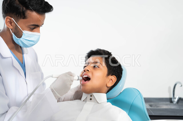 ولد يجلس في العيادة السنية يتلقى العلاج اللازم، طبيب أسنان عربي خليجي سعودي يرتدي المعطف والقفازات والكمامة الطبية يمسك بيده أداة حفر السن وإزالة التسوس، زيارة العيادات الصحية واتخاذ الإجراءات اللازمة
