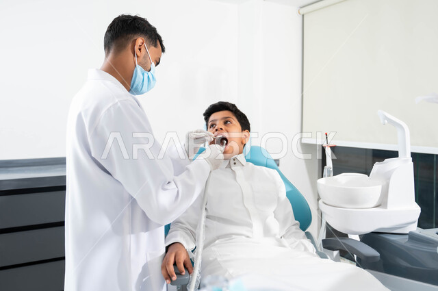 زيارة العيادات الصحية واتخاذ الإجراءات اللازمة، طبيب أسنان عربي خليجي سعودي يرتدي المعطف والقفازات والكمامة الطبية يمسك بيده أداة حفر السن وإزالة التسوس، ولد يجلس في العيادة السنية يتلقى العلاج اللازم