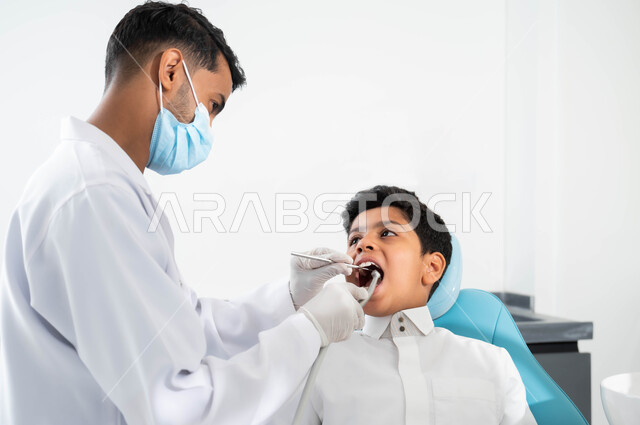 زيارة العيادات الصحية واتخاذ الإجراءات اللازمة، صورة مقربة لطبيب أسنان عربي خليجي سعودي يرتدي القفازات الطبية يمسك بيده أداة حفر السن وإزالة التسوس، ولد سعودي يجلس في العيادة السنية ويفتح فمه لمعالجة النخور و يتلقى العلاج اللازم, التجهيز لتركيب الحشوات