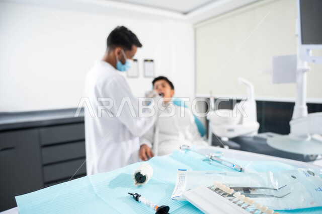 صورة مقربة للمعدات اللازمة لتركيب الحشوات ,زيارة العيادات الصحية واتخاذ الإجراءات اللازمة، طبيب أسنان عربي خليجي سعودي يرتدي القفازات الطبية يمسك بيده أداة حفر السن وإزالة التسوس، ولد سعودي يجلس في العيادة السنية ويفتح فمه لمعالجة النخور و يتلقى العلاج اللازم