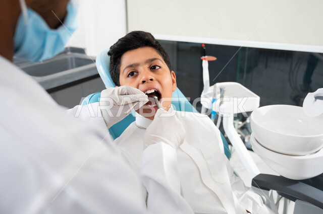 التجهيز لتركيب الحشوات ,زيارة العيادات الصحية واتخاذ الإجراءات اللازمة، صورة مقربة من  الخلف لطبيب أسنان عربي خليجي سعودي يرتدي القفازات الطبية يمسك بيده أداة حفر السن وإزالة التسوس، ولد سعودي يجلس في العيادة السنية ويفتح فمه لمعالجة النخور و يتلقى العلاج اللازم