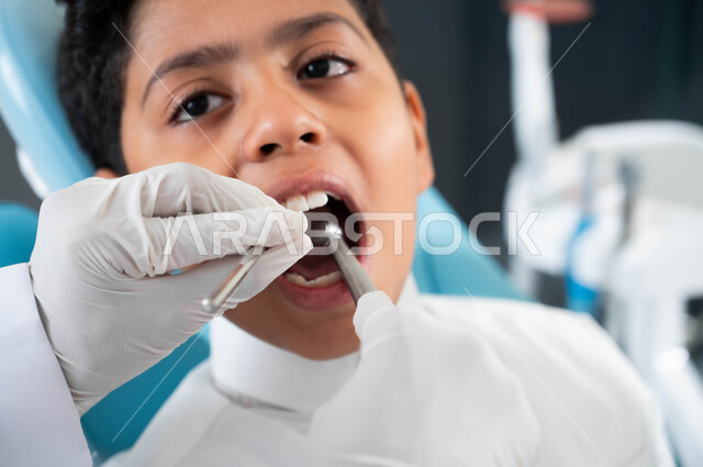 زيارة العيادات الصحية واتخاذ الإجراءات اللازمة، أيدي طبيب أسنان عربي خليجي سعودي يرتدي القفازات الطبية يمسك بيده أداة حفر السن وإزالة التسوس، معالجة النخور و التجهيز لتركيب الحشوات , صورة مقربة لولد سعودي يجلس في العيادة السنية ويفتح فمه ليتلقى العلاج اللازم