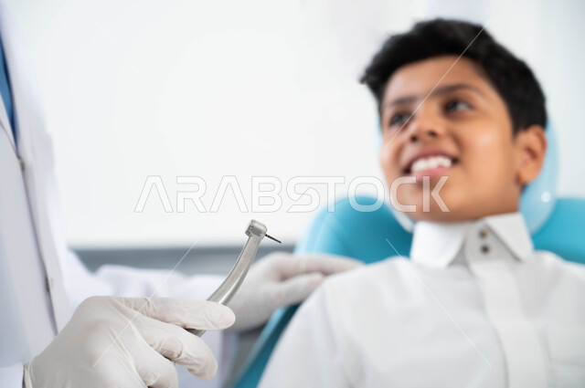 معالجة النخور و التجهيز لتركيب الحشوات ,زيارة العيادات الصحية واتخاذ الإجراءات اللازمة، أيدي طبيب أسنان عربي خليجي سعودي يرتدي القفازات الطبية يمسك بيده أداة حفر السن وإزالة التسوس، صورة مقربة لولد سعودي يجلس في العيادة السنية يتلقى العلاج اللازم