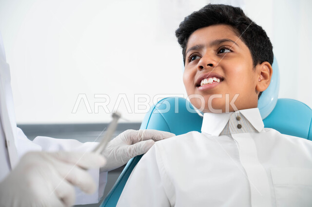 معالجة النخور والتجهيز لتركيب الحشوات ,زيارة العيادات الصحية واتخاذ الإجراءات اللازمة، صورة مقربة لأيدي طبيب أسنان عربي خليجي سعودي يرتدي القفازات الطبية يمسك بيده أداة حفر السن وإزالة التسوس، ولد يجلس في العيادة السنية يتلقى العلاج اللازم