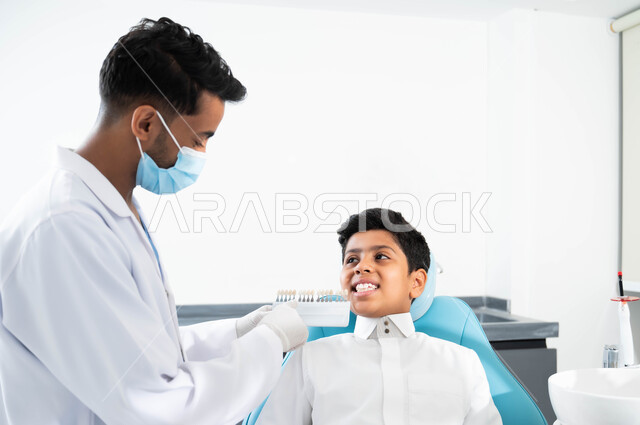 الحصول على ابتسامة مثالية ,تقديم الرعاية للأطفال في المراكز التخصصية، ولد عربي خليجي سعودي يجلس في العيادة السنية يختار اللون المناسب من قشور الفينير, صورة مقربة لطبيب عربي خليجي سعودي يرتدي المعطف القفازات و الكمامة الطبية تمسك بيدها دليل الألوان