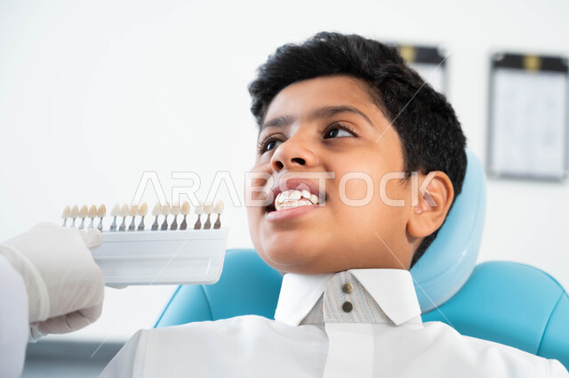 ترميم أسنان المريض للحصول على ابتسامة مثالية ,تقديم الرعاية للأطفال في المراكز التخصصية، ولد عربي خليجي سعودي يجلس في العيادة السنية يختار اللون المناسب من قشور الفينير, صورة مقربة لأيدي طبيبة عربية خليجية سعودية ترتدي القفازات الطبية تمسك بيدها دليل الألوان