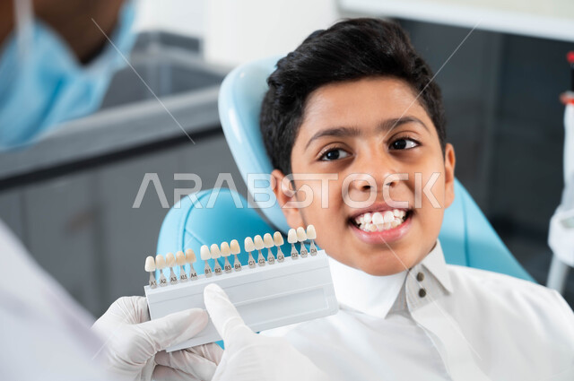 الحصول على ابتسامة مثالية ,تقديم الرعاية للأطفال في المراكز التخصصية، ولد عربي خليجي سعودي يجلس في العيادة السنية يختار اللون المناسب من قشور الفينير, صورة مقربة لأيدي طبيبة عربية خليجية سعودية ترتدي القفازات الطبية تمسك بيدها دليل الألوان لترميم أسنان المريض