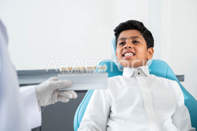 تقديم الرعاية للأطفال في المراكز التخصصية، صورة مقربة لولد عربي خليجي سعودي يجلس في العيادة السنية يختار اللون المناسب من قشور الفينير, أيدي طبيبة عربية خليجية سعودية ترتدي القفازات الطبية تمسك بيدها دليل الألوان لترميم أسنان المريض, الحصول على ابتسامة مثالية