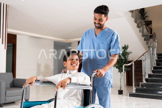 Paying attention to the psychological aspect of people of determination, Saudi companies specializing in home nursing, taking care of people with special needs, a Saudi Gulf Arab nurse wearing a work uniform who helps a Saudi boy sitting in a wheelchair move around inside the home, providing equipment that helps with movement.