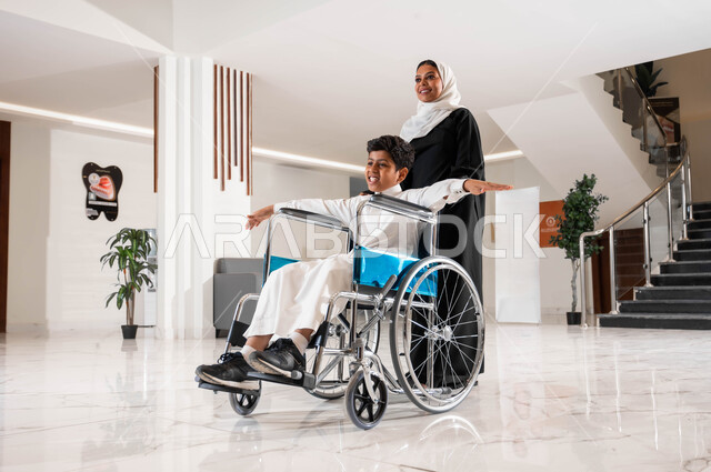 تأمين معدات تساعد على الحركة ,الاهتمام بالجانب النفسي لأصحاب الهمم, رعاية الأبناء والاعتناء بذوي الاحتياجات الخاصة, أم عربية سعودية خليجية محجبة تقوم بمساعدة ابنها في التنقل داخل المنزل, ولد سعودي مقعد يجلس على كرسي متحرك يمد يديه جانبا بإيماءات السرور
