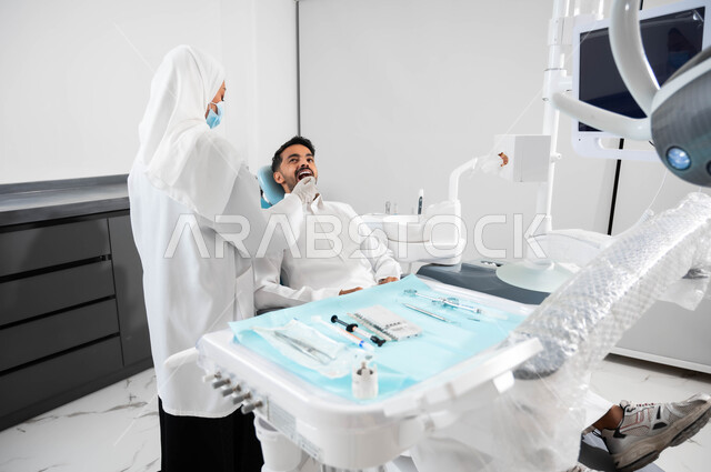 استخدام المعدات اللازمة لجراحة الفم، صورة من الخلف لطبيبة عربية خليجية سعودية تقوم بفحص أسنان المريض و تشخيص المرض، شاب عربي خليجي سعودي يرتدي الزي التقليدي و يجلس في العيادة السنية يقوم بإجراء الفحوصات للعناية بالصحة الفموية, معالجة التسوس وتركيب الحشوات