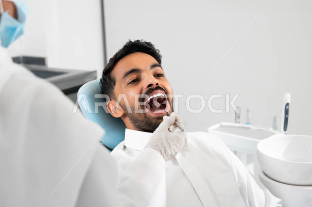 معالجة التسوس و النخر, استخدام الادوات اللازمة لإجراء الفحوصات الطبيه ,شاب عربي خليجي سعودي يرتدي الزي التقليدي يجلس في العيادة السنية ويفتح فمه لتشخيص المرض, صورة مقربة لطبيبة عربية خليجيه سعودية ترتدي المعطف و القفازات والكمامة الطبية تقوم بفحص أسنان المريض