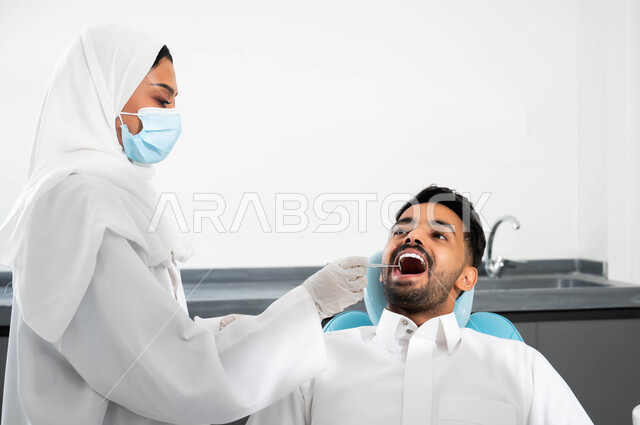 معالجة النخر والتسوس, استخدام الادوات اللازمة لإجراء الفحوصات الطبيه ,شاب عربي خليجي سعودي يرتدي الزي التقليدي يجلس في العيادة السنية ويفتح فمه لتشخيص المرض, صورة مقربة لطبيبة عربية خليجيه سعودية ترتدي المعطف و القفازات والكمامة الطبية تقوم بفحص أسنان المريض