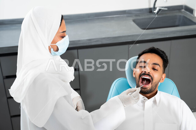 Using the necessary tools to conduct medical examinations, treat caries and caries, a Saudi Gulf Arab young man wearing traditional clothing sits in the dental clinic and opens his mouth to diagnose the disease, a close-up picture of a Saudi Gulf Arab doctor wearing a coat, gloves and a medical mask examining the patient’s teeth