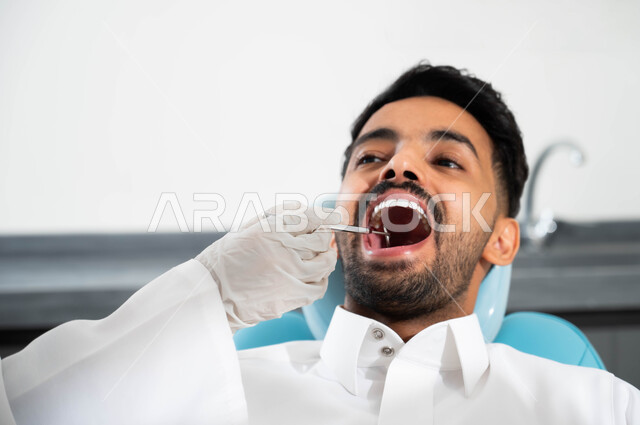 معالجة النخور وتركيب الحشوات، طبيبة عربية خليجية سعودية تقوم بفحص أسنان المريض و تشخيص المرض، إجراء الفحوصات الروتينية ,صورة مقربة لشاب عربي خليجي سعودي يرتدي الزي التقليدي و يجلس في العيادة السنية يقوم بالزيارة الدورية للاطمئنان على الصحة الفموية