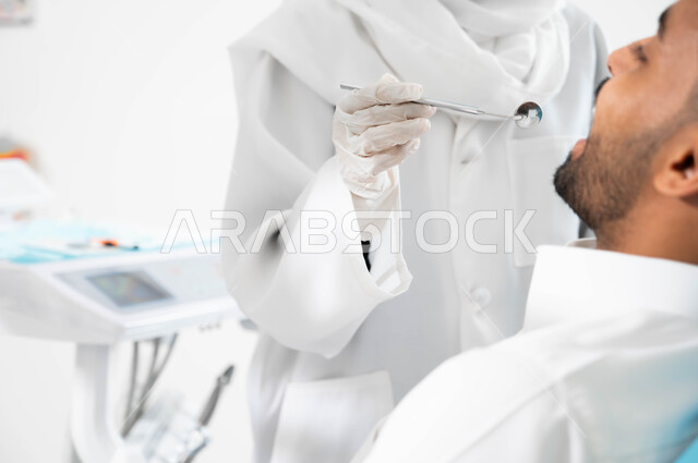 Conducting routine examinations, treating caries and installing fillings, a Saudi Gulf Arab doctor examines the patient’s teeth and diagnoses the disease, a close-up picture from the side of a Saudi Gulf Arab young man wearing traditional clothing and sitting in the dental clinic making a periodic visit to check on his oral health.