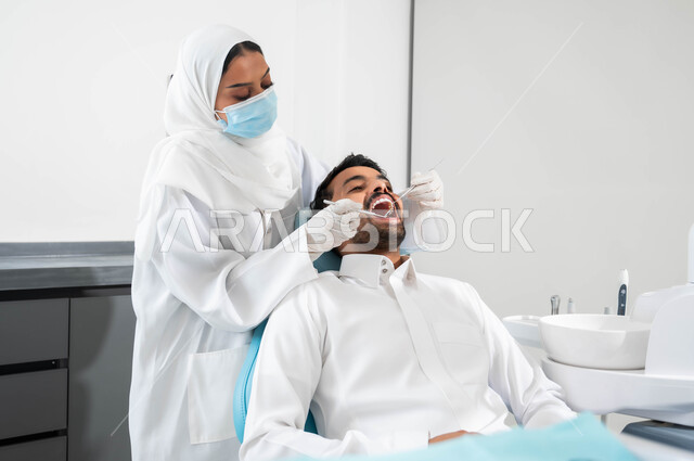 استخدام المعدات والأدوات اللازمة لجراحة الفم, معالجة النخور وتركيب الحشوات، طبيبة عربية خليجية سعودية تقوم بفحص أسنان المريض و تشخيص المرض، شاب عربي خليجي سعودي يرتدي الزي التقليدي و يجلس في العيادة السنية يقوم بإجراء الفحوصات للعناية بالصحة الفموية