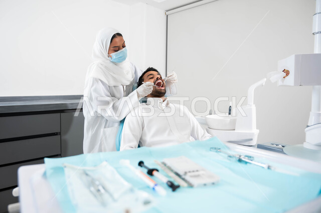 معالجة النخور وتركيب الحشوات، المعدات والأدوات اللازمة لجراحة الفم, طبيبة عربية خليجية سعودية تقوم بفحص أسنان المريض و تشخيص المرض، شاب عربي خليجي سعودي يرتدي الزي التقليدي و يجلس في العيادة السنية يقوم بإجراء الفحوصات للعناية بالصحة الفموية