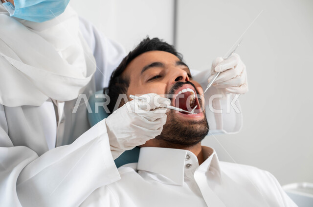 معالجة النخر والتسوس, استخدام الادوات والمعدات اللازمة لإجراء الفحوصات الطبيه, صورة مقربة لشاب عربي خليجي سعودي يرتدي الزي التقليدي يجلس في العيادة السنية ويفتح فمه لتشخيص المرض, ايدي طبيبة عربية خليجيه سعودية ترتدي القفازات الطبية تقوم بفحص أسنان المريض