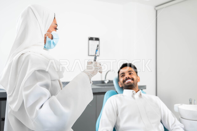 معالجة التسوس والنخور ,الاستعداد لحقن البنج الموضعي لتخفيف الشعور بالألم ,تقديم الرعاية الصحية في المراكز التخصصية، رجل عربي خليجي سعودي يجلس في العيادة السنية بإيماءات السرور,  صورة مقربة لطبيبة عربية خليجية سعودية ترتدي القفازات الطبية تمسك بيدها ابرة المخدر