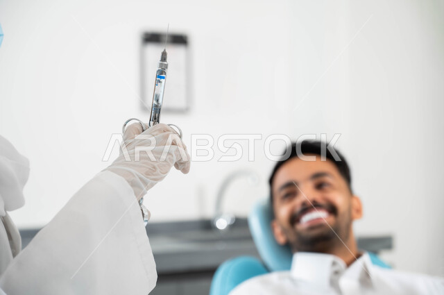 معالجة النخور والتسوس ,الاستعداد لحقن البنج الموضعي لتخفيف الشعور بالألم ,تقديم الرعاية الصحية في المراكز التخصصية، صورة مقربة لرجل عربي خليجي سعودي يجلس في العيادة السنية بإيماءات السرور, يد طبيبة عربية خليجية سعودية ترتدي القفازات الطبية تمسك بيدها ابرة المخدر