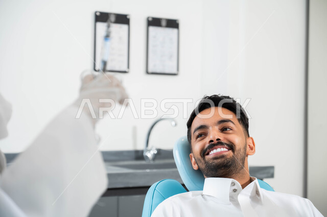 الاستعداد لحقن البنج الموضعي لتخفيف الشعور بالألم ,تقديم الرعاية الصحية في المراكز التخصصية، صورة مقربة لرجل عربي خليجي سعودي يجلس في العيادة السنية بإيماءات السرور, يد طبيبة عربية خليجية سعودية ترتدي القفازات الطبية تمسك بيدها ابرة المخدر, معالجة النخور والتسوس