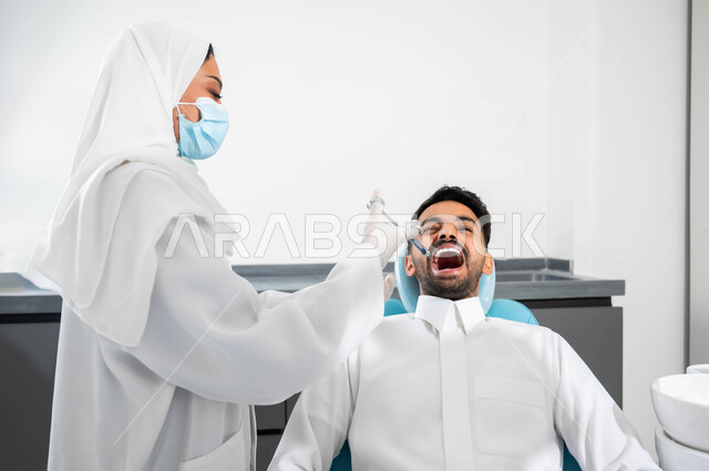 معالجة الأسنان بالحشوات التجميلية، مفهوم تخفيف الشعور بالألم، طبيبة أسنان عربية خليجية سعودية ترتدي القفازات الطبية تمسك بيدها بحقن البنج الموضعي باستخدام الابرة، شاب عربي خليجي سعودي يرتدي الزي التقليدي يجلس في عيادة سنية يتلقى العلاج اللازم لفمه
