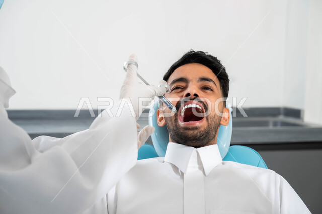 مفهوم تخفيف الشعور بالألم، ترميم الأسنان بالحشوات التجميلية، طبيبة أسنان عربية خليجية سعودية ترتدي القفازات الطبية تمسك بيدها بحقن البنج الموضعي باستخدام الابرة، صورة مقربة لشاب عربي خليجي سعودي يرتدي الزي التقليدي يجلس في عيادة سنية يتلقى العلاج اللازم لفمه