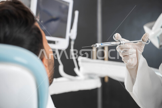 حقن البنج الموضعي لتخفيف الشعور بالألم، ترميم الأسنان بالحشوات التجميلية، طبيبة أسنان عربية خليجية سعودية ترتدي القفازات الطبية تمسك بيدها إبرة التخدير، صورة مقربة من الخلف لشاب عربي خليجي سعودي يرتدي الزي التقليدي يجلس في عيادة سنية يتلقى العلاج اللازم لفمه