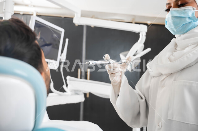 ترميم الأسنان بالحشوات التجميلية، صورة مقربة لطبيبة أسنان عربية خليجية سعودية ترتدي القفازات الطبية تمسك بيدها إبرة التخدير، شاب عربي خليجي سعودي يرتدي الزي التقليدي يجلس في عيادة سنية يتلقى العلاج اللازم لفمه, حقن البنج الموضعي لتخفيف الإحساس بالألم