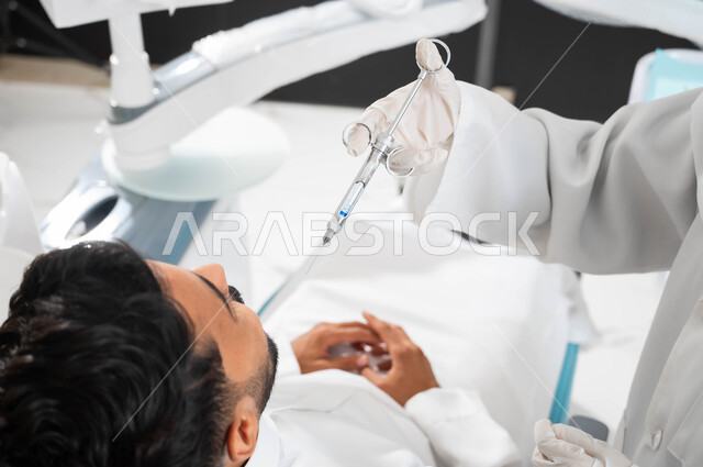 ترميم الأسنان بالحشوات التجميلية، طبيبة أسنان عربية خليجية سعودية ترتدي القفازات الطبية تمسك بيدها إبرة التخدير، حقن البنج الموضعي لتخفيف الإحساس بالألم، صورة مقربة من الخلف لشاب عربي خليجي سعودي يرتدي الزي التقليدي يجلس في عيادة سنية يتلقى العلاج اللازم لفمه