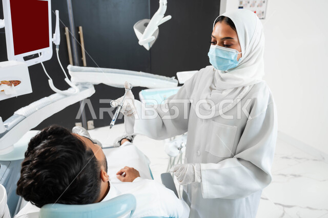 الادوات اللازمة لحقن البنج الموضعي لتخفيف الشعور بالألم، ترميم الأسنان بالحشوات التجميلية، طبيبة أسنان عربية خليجية سعودية ترتدي القفازات الطبية تمسك بيدها إبرة التخدير، شاب عربي خليجي سعودي يرتدي الزي التقليدي يجلس في عيادة سنية يتلقى العلاج اللازم لفمه