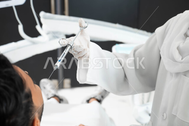 ترميم الأسنان بالحشوات التجميلية، طبيبة أسنان عربية خليجية سعودية ترتدي القفازات الطبية تمسك بيدها إبرة التخدير، حقن البنج الموضعي لتخفيف الإحساس بالألم، صورة مقربة من الخلف لشاب عربي خليجي سعودي يرتدي الزي التقليدي يجلس في عيادة سنية يتلقى العلاج اللازم لفمه