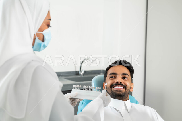 تقديم الخدمات الصحية في المراكز التخصصية، صورة مقربة من الخلف لطبيبة أسنان عربية خليجية سعودية ترتدي المعطف والكمامة الطبية تمسك بيدها دليل الألوان لترميم أسنان المريض، رجل عربي خليجي سعودي يجلس في العيادة السنية يتلقى العلاج اللازم للحصول على ابتسامة مثالية