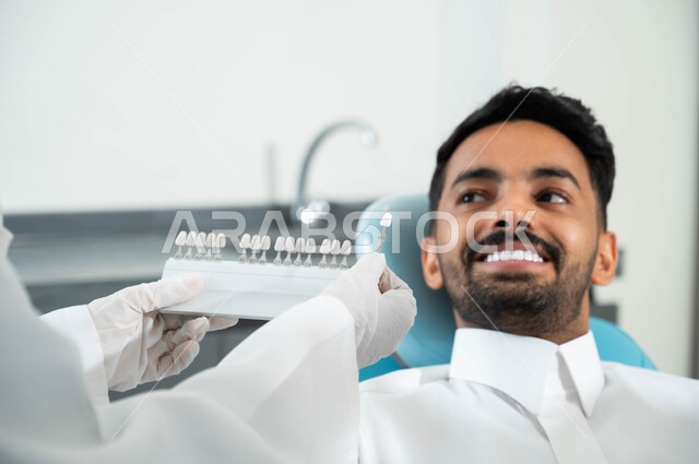 اختيار اللون المناسب من قشور الفينير ,الحصول على ابتسامة مثالية ,تقديم الرعاية الصحية في المراكز التخصصية، صورة مقربة لأيدي طبيبة عربية خليجية سعودية ترتدي القفازات الطبية تمسك بيدها دليل الألوان لترميم أسنان المريض، رجل عربي خليجي سعودي يجلس في العيادة السنية
