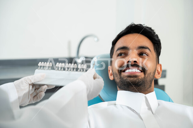 اختيار اللون المناسب من قشور الفينير ,الحصول على ابتسامة مثالية ,تقديم الرعاية الصحية في المراكز التخصصية، صورة مقربة لأيدي طبيبة عربية خليجية سعودية ترتدي القفازات الطبية تمسك بيدها دليل الألوان لترميم أسنان المريض، رجل عربي خليجي سعودي يجلس في العيادة السنية