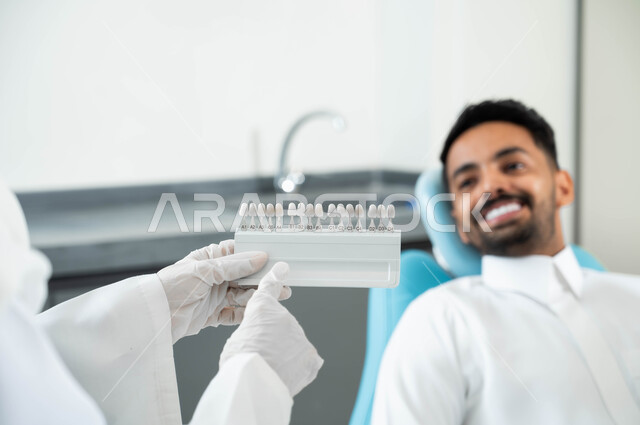 الحصول على ابتسامة مثالية ,تقديم الرعاية الصحية في المراكز التخصصية، صورة مقربة لأيدي طبيبة عربية خليجية سعودية ترتدي القفازات الطبية تمسك بيدها دليل الألوان لترميم أسنان المريض، رجل عربي خليجي سعودي يجلس في العيادة السنية يختار اللون المناسب من قشور الفينير