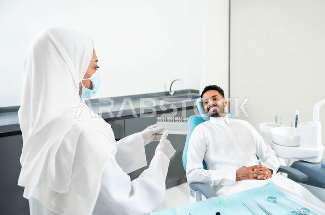 Providing health care in specialized centers, a picture from the back of a Saudi Gulf Arab dentist wearing a coat and a medical mask, holding in her hand a color guide for restoring the patient’s teeth. A Saudi Gulf Arab man sitting in the dental clinic receiving the necessary treatment to obtain a perfect smile.