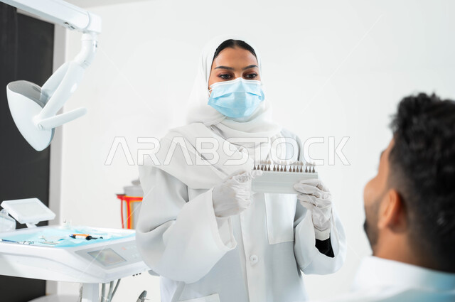 Providing health care and services in specialized centers. A Saudi Gulf Arab dentist wearing a coat, gloves and a medical mask holds in her hand a color guide for restoring the patient’s teeth. A Saudi Gulf Arab man sits in the dental clinic receiving the necessary treatment to obtain a perfect smile.