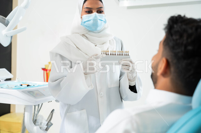 Providing services and health care in specialized centers. A Saudi Gulf Arab dentist wearing a coat, gloves, and a medical mask holds in her hand a color guide for restoring the patient’s teeth. A Saudi Gulf Arab man sits in the dental clinic receiving the necessary treatment to obtain a perfect smile.