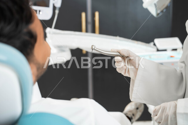 المعدات اللازمة لإجراء الفحوصات الطبية, التجهبز لتركيب الحشوات, صورة مقربة ليد طبيبة عربية خليجيه سعودية ترتدي القفازات الطبية تحمل آلة الحفر لمعالجة التسوس في أسنان المريض, شاب عربي خليجي سعودي يقوم بالزيارة الدورية للعيادة السنية للاهتمام بصحة الفم
