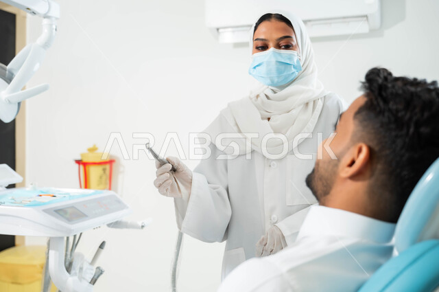 The equipment needed to conduct medical examinations, the therapeutic methods used for oral care, a close-up picture of a young Saudi Gulf Arab man sitting in the dental clinic, a Saudi Gulf Arab doctor wearing gloves and a medical mask, holding a drilling machine in her hand and preparing to begin treating caries in the patient’s teeth.