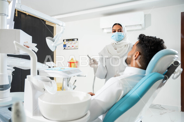 الأساليب العلاجية المتبعة للعناية بالفم, شاب عربي خليجي سعودي يجلس في العيادة السنية, طبيبة عربية خليجيه سعودية ترتدي القفازات والمعطف و الكمامة الطبية تحمل بيدها آلة الحفر وتستعد لبدء معالجة التسوس في أسنان المريض, المعدات اللازمة لإجراء الفحوصات الطبية