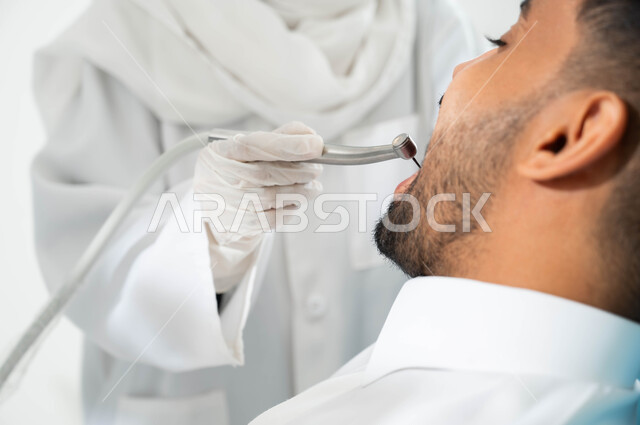 التجهبز لتركيب الحشوات, صورة مقربة لشاب عربي خليجي سعودي يجلس في العيادة السنية ويفتح فمه لتشخيص المرض, ايدي طبيبة عربية خليجيه سعودية ترتدي القفازات الطبية تستخدم آلة الحفر لمعالجة التسوس في أسنان المريض, المعدات اللازمة لإجراء الفحوصات الطبية